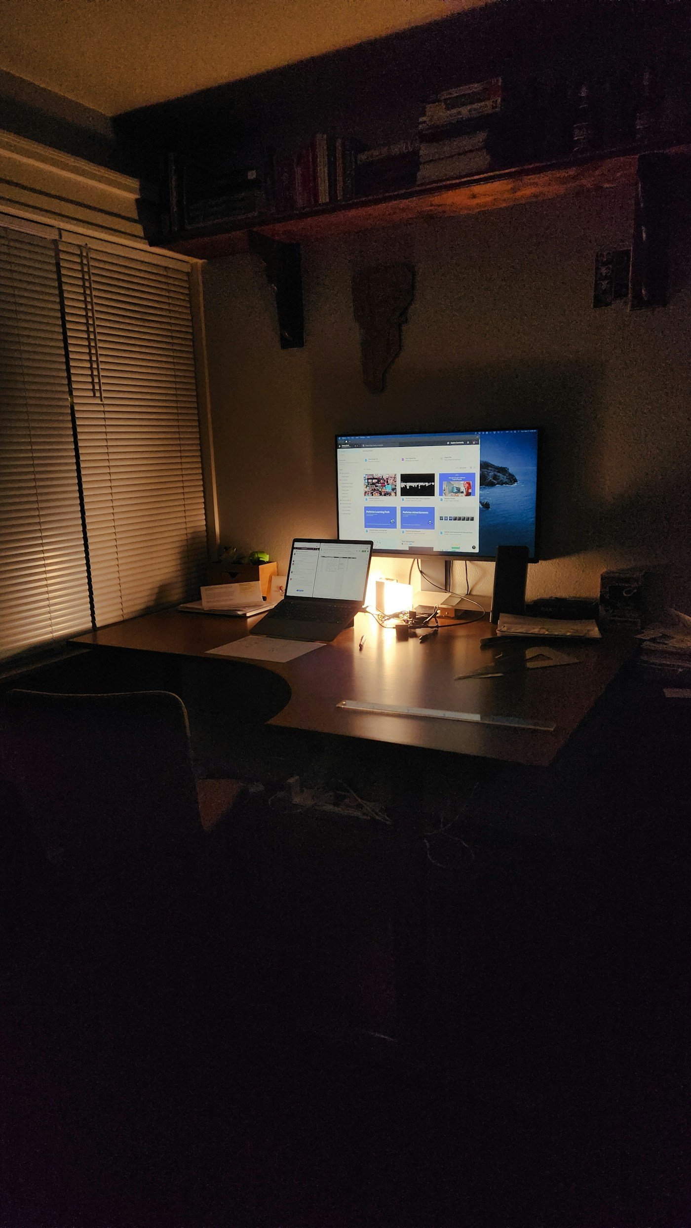 A dim room with a monitor and laptop glowing on a desk lit only by a small warm lamp