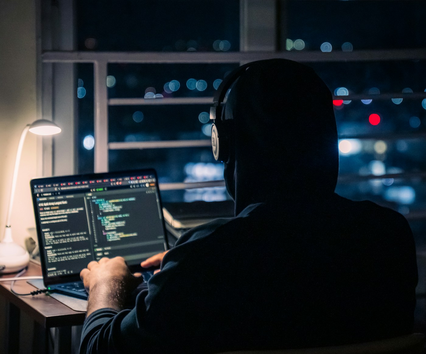 Silhouette of a builder at a laptop at night, city lights blurred in the window behind